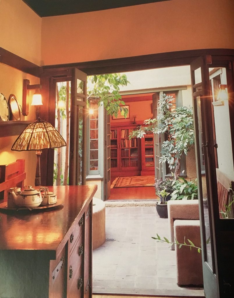 View through open French doors from a tearoom at the Duncan-Irwin House into the central courtyard, with a Tiffany-style lamp on a built-in cabinet in the foreground and the library visible across the courtyard beyond.