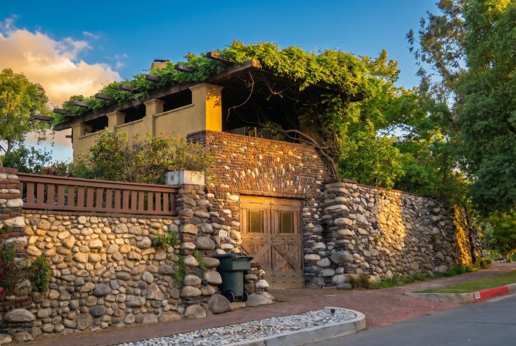 A wisteria-draped pergola above a river-cobble wall with a wood carriage door, Arroyo Terrace beginning to curve south in front of Charles Greene's family home.