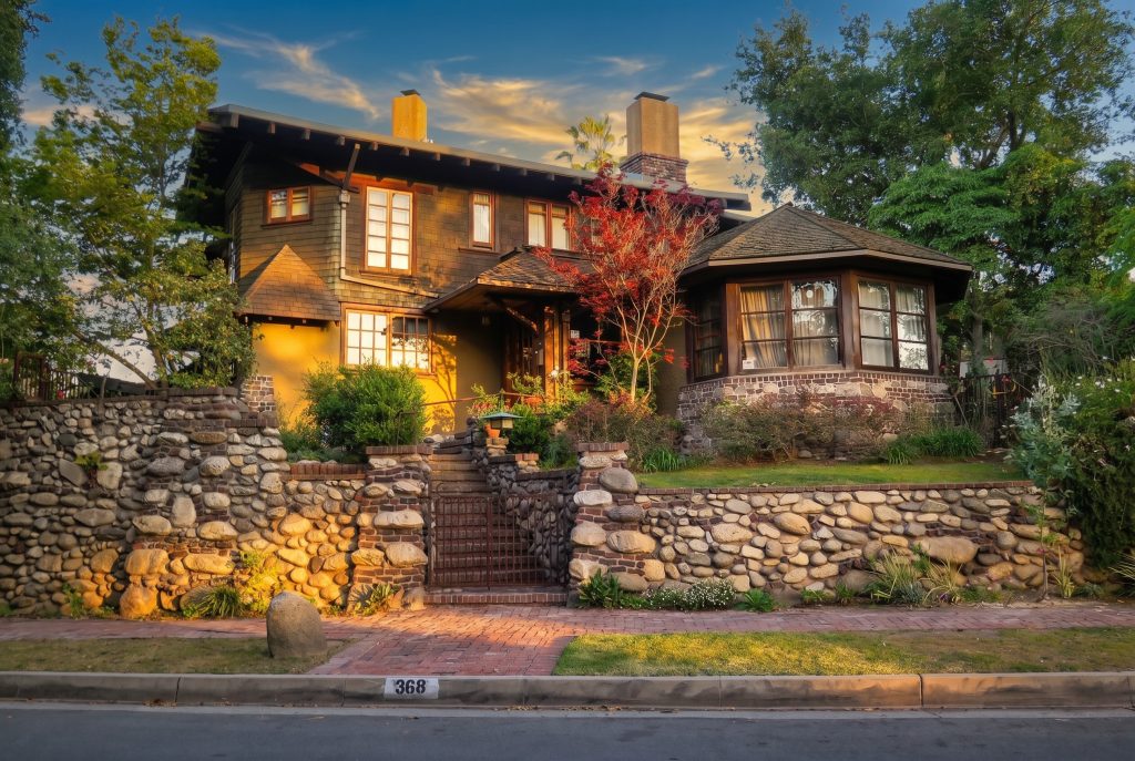 The Charles Greene House with a bay window and a red maple, fronted by a river-cobble wall with brick-trimmed piers and a central iron gate.