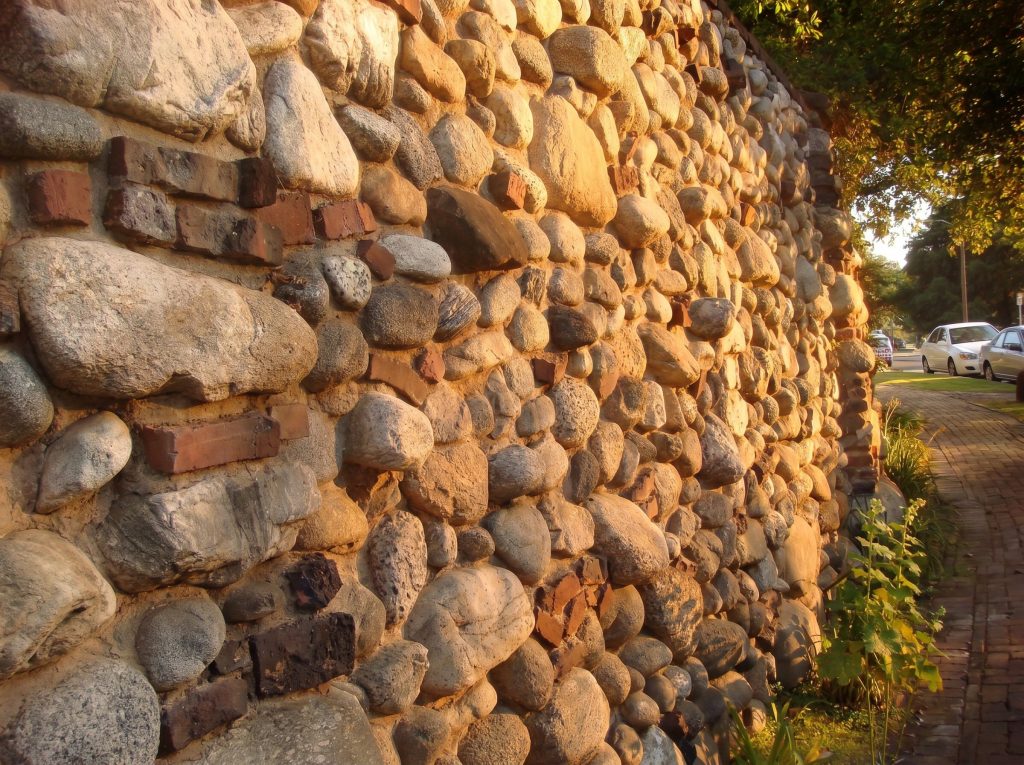 Close view of the wall surface as it rounds the corner past Charles Greene's house: tightly fitted rounded granite cobbles with occasional red and dark brick courses threaded between them, lit by low sun.