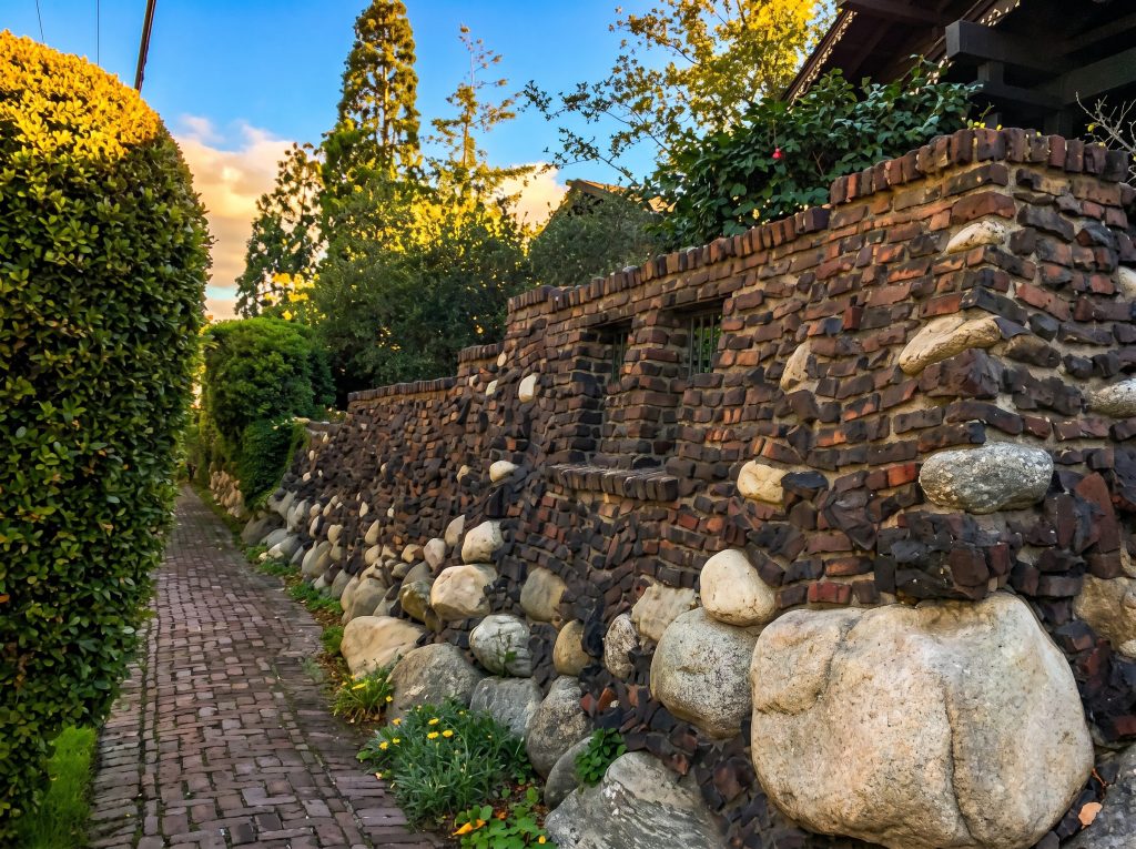 A brick sidewalk running beside a boulder-and-clinker-brick wall with a rounded brick cap and small barred openings, a clipped hedge opposite, under a sunset sky, looking east in front of The van Rossem-Neil House.