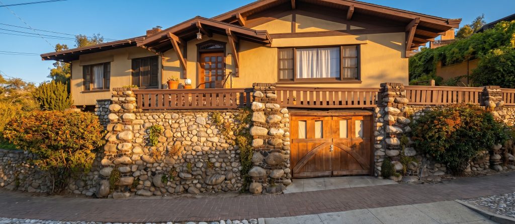 The White Sister's House with a wood-railed porch rising above a street wall of rounded granite cobbles, a wood plank gate set into the wall between cobble piers, in golden light.