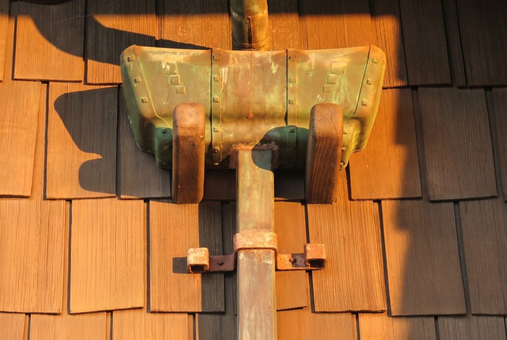 Close-up frontal view of a copper leader head on the Gamble House, showing the three riveted panels of the box collector with embossed square detailing, the green and warm orange patina of weathered copper, two shaped wooden corbel brackets supporting the box from below against the shingled wall, the round copper inlet pipe descending into the top of the box, and the round outlet pipe continuing below with a simple rusted iron strap securing it to the wall.