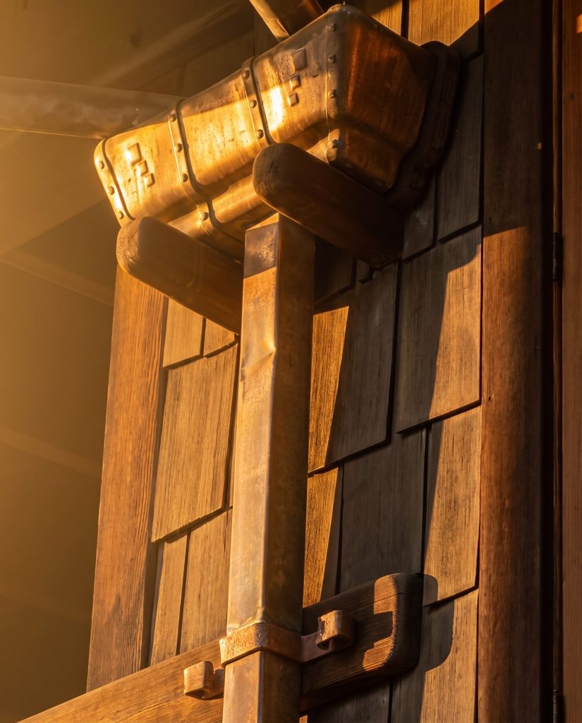 Side three-quarter view of a copper leader head on the Gamble House in late-afternoon golden light, showing the multi-paneled form of the collector with three connected box sections in profile, two shaped wooden corbel brackets supporting the assembly against the shingled wall, the round copper inlet entering at the top and the round outlet descending past a horizontal wooden strap and a smaller wooden block at the lower trim.
