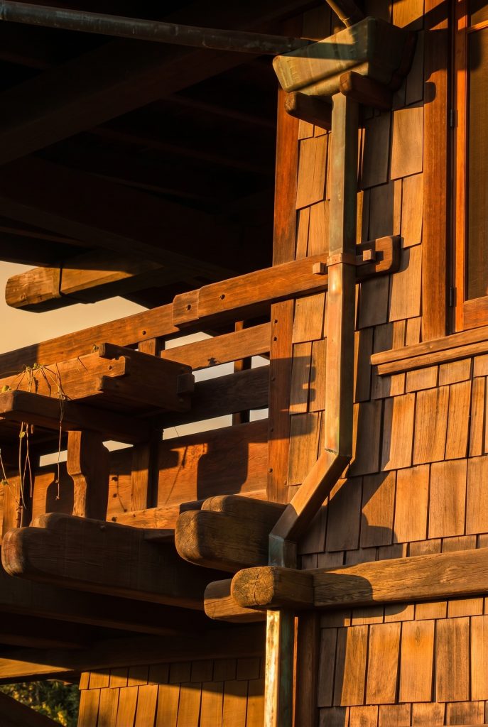 Late-afternoon side view at a corner of the Gamble House, showing a copper downspout running vertically between the wood-shingled wall and the structural timber assembly of the projecting eave and second-story sleeping porch, with shaped beam ends and outriggers visible at multiple stacked levels, a small copper collector at the top of frame, and the round pipe threading past the brackets and sleeping porch rails on its way down the wall.