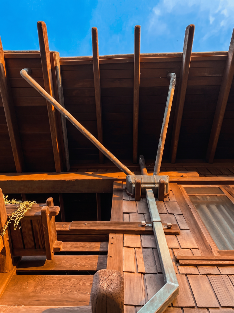 Looking up at the underside of the eave at the Gamble House, showing the deep timber roof structure with rafters and dropped beams overhead, two angled wooden brace members descending to a copper box leader head fastened against the wood-shingled wall, the round copper downspout running vertically away from the leader, a wood-railed balcony with shaped end-pieces in the foreground, and the post-end visible at lower right.