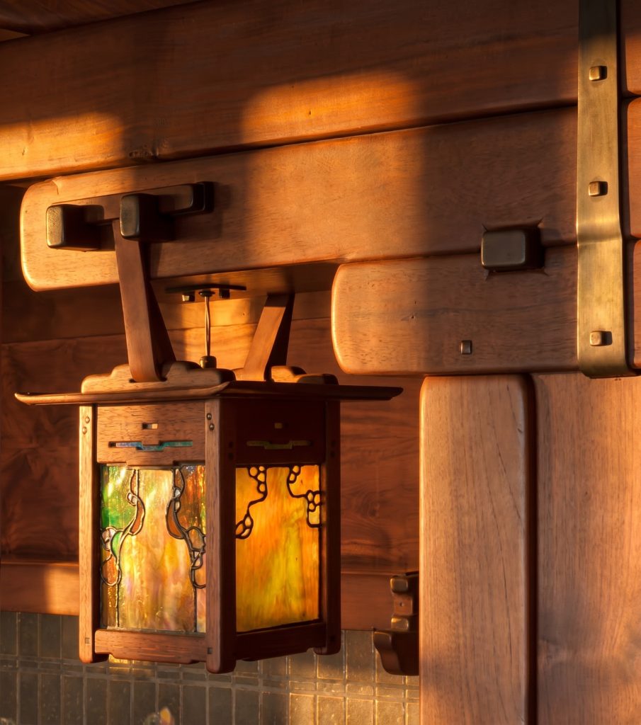 Close view of a Gamble House lantern suspended from a leather strap that loops over a projecting forged-iron bracket fixed to the wooden post, the strap's grain and edges clearly visible, the square art-glass lantern hanging below