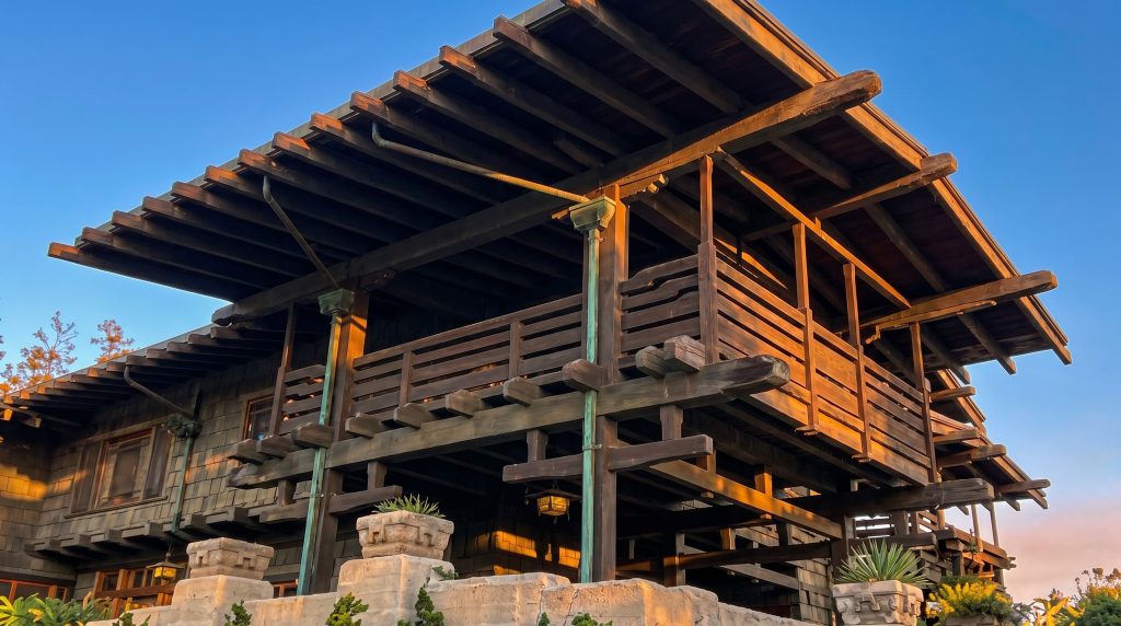Upward corner view of a Gamble House sleeping porch, showing the great cantilevered eaves, exposed beams with rounded ends, verdigris copper posts and downspouts, and the open timber-railed porch rising above a boulder-and-clinker terrace base at sunset.
