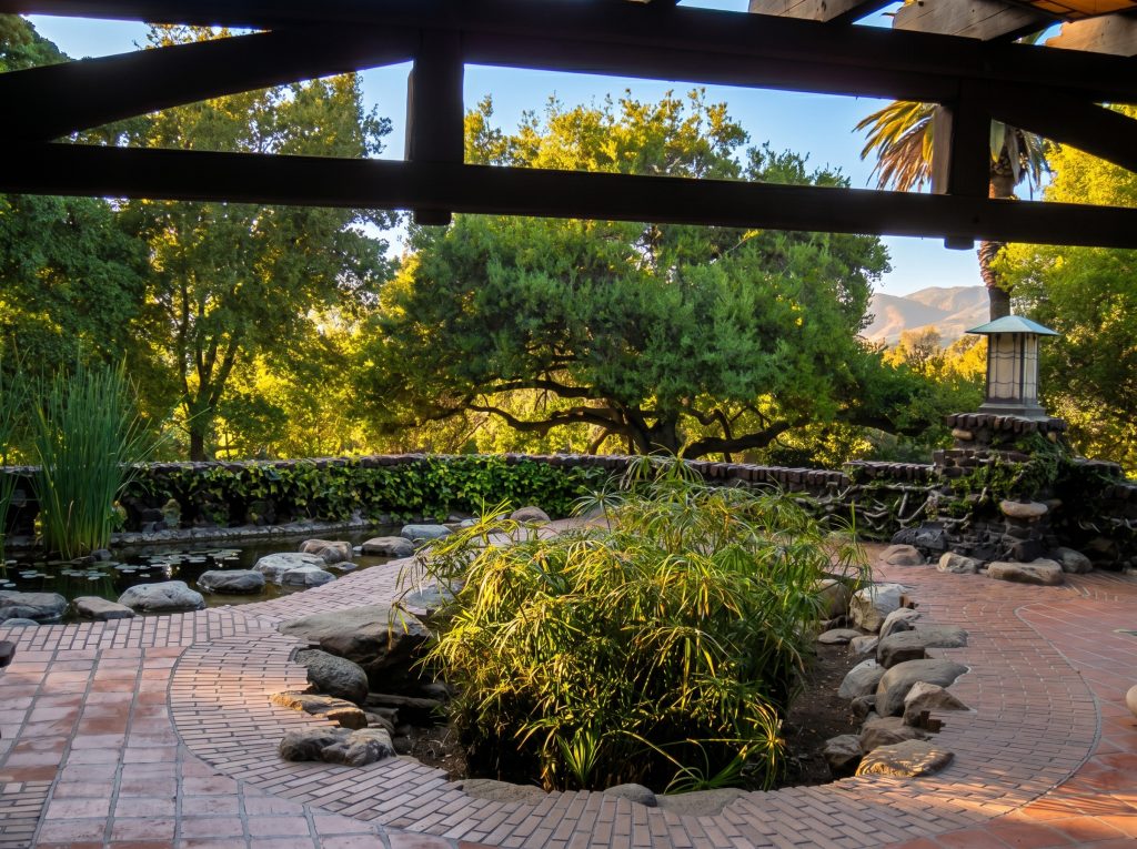 A view from beneath the heavy timber trellis at the Gamble House, looking out over a brick terrace to the stone-walled garden pond, a planted island of umbrella sedge, a hanging metal lantern, spreading oak trees, and mountains in the distance under a clear sky.
