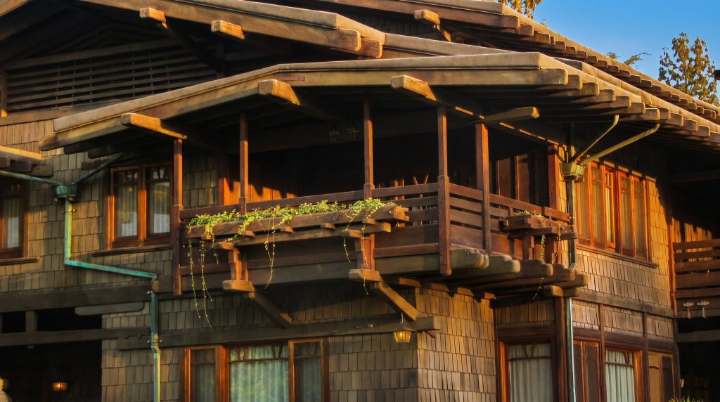 A second-floor sleeping porch on the David B. Gamble House, its open timber frame and horizontal rail projecting beneath deep overhanging eaves and exposed rafters, with a planter box of trailing greenery and shingled walls glowing in late-afternoon light.