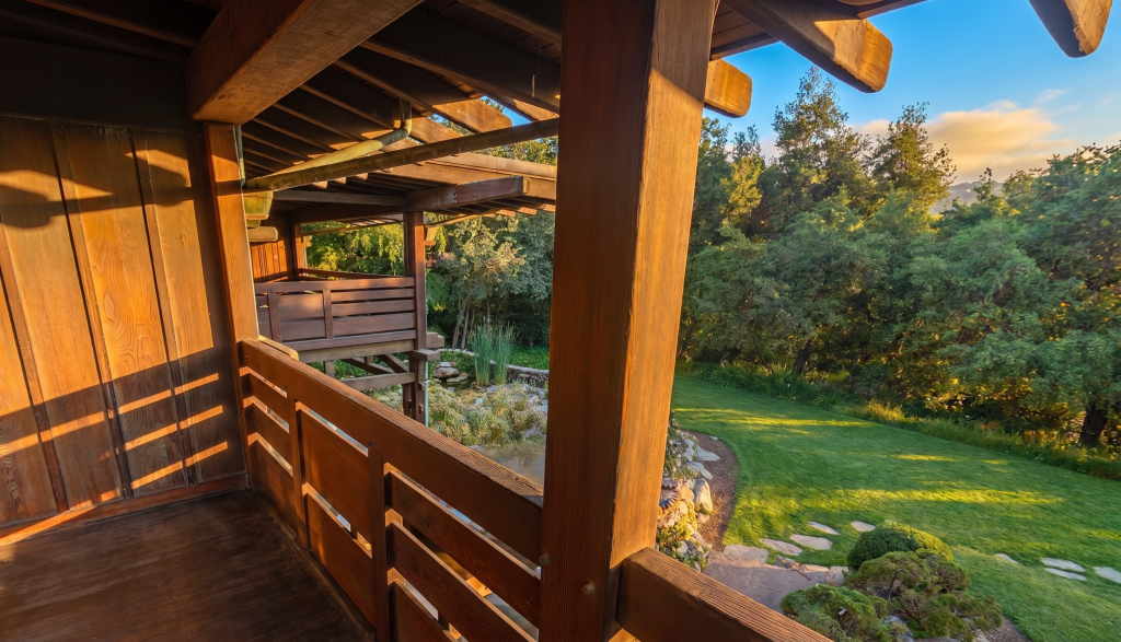 The view from a Gamble House sleeping porch, looking past a timber post and rail down to the rear terrace and the green backyard beyond — lawn, trees, a stone path, and a rock-and-water garden in golden light.