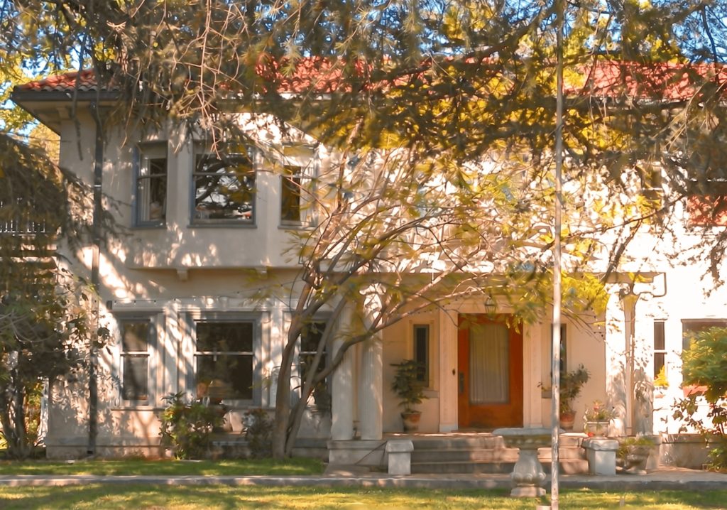 The James Swan House today, seen from the street through mature trees: a two-story stuccoed residence with a red clay-tile roof, a columned entrance portico sheltering a wood front door, and a projecting second-floor bay window, in dappled afternoon light.
