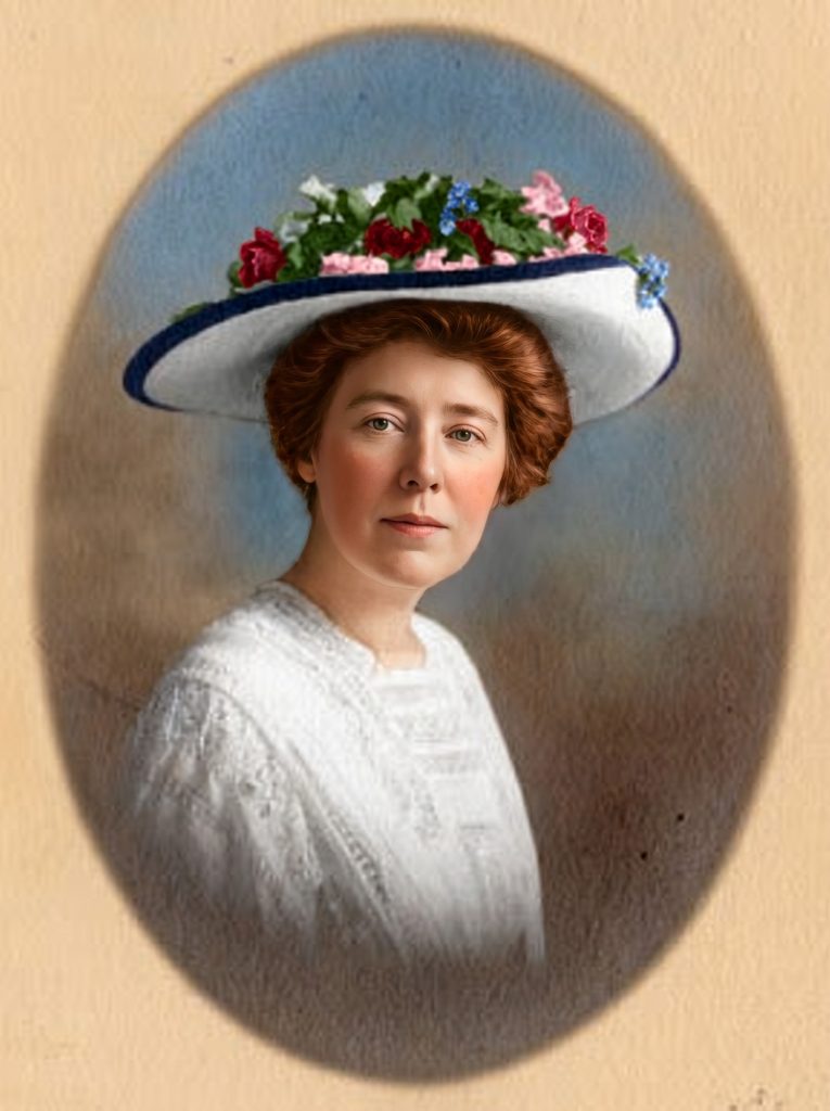Colorized oval-framed portrait of a woman in a wide white hat trimmed with red and pink roses and blue flowers, wearing a white dress.