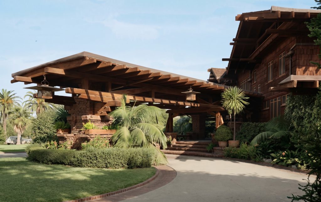 Side view of the Robert R. Blacker House porte-cochère, its stepped timber roof beams cantilevering outward like a ship's prow over the driveway, two lanterns hanging beneath, brick piers below, with the shingled house at right and palms and clipped hedges lining the drive.