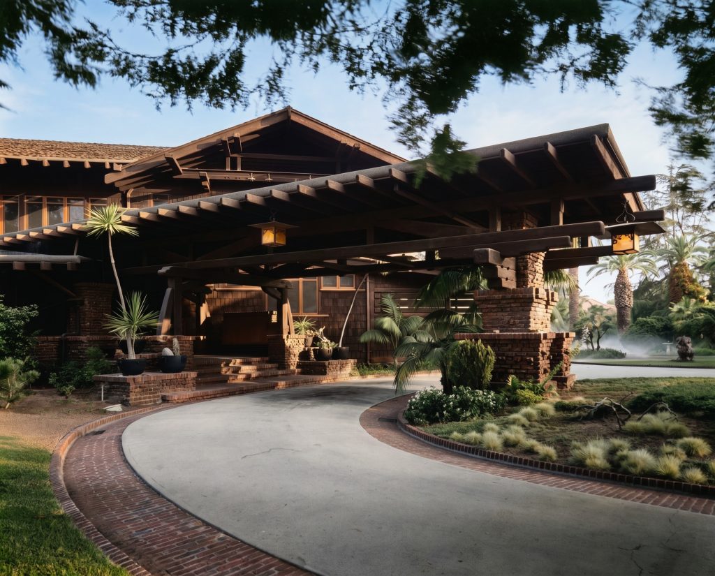 The porte-cochère of the Robert R. Blacker House, a broad cantilevered timber roof carried on clinker-brick piers reaching out over a curving driveway, an iridescent art-glass lantern hanging beneath it, with the shingled house and tropical plantings behind in low warm sunlight.