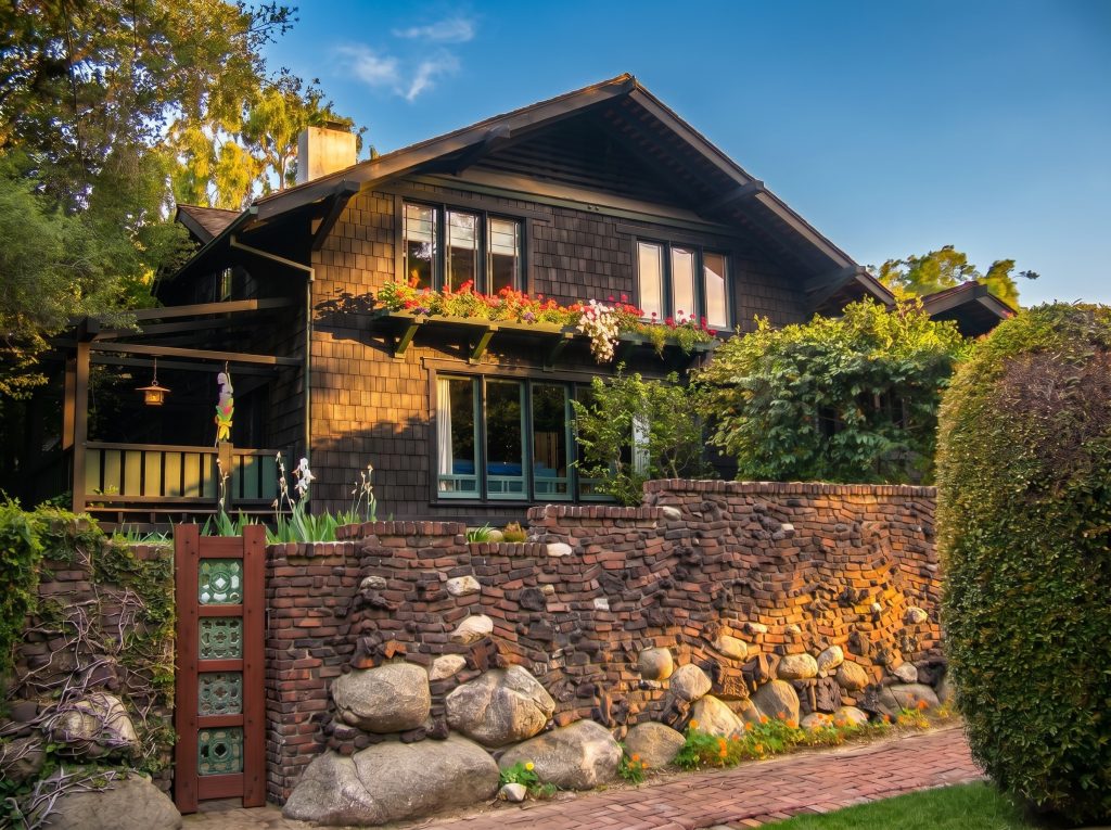 A two-story brown-shingled Craftsman house at golden hour behind a wall of clinker brick and river boulders, with red geraniums in a window box and a wood-framed green tile panel set into the wall at left.