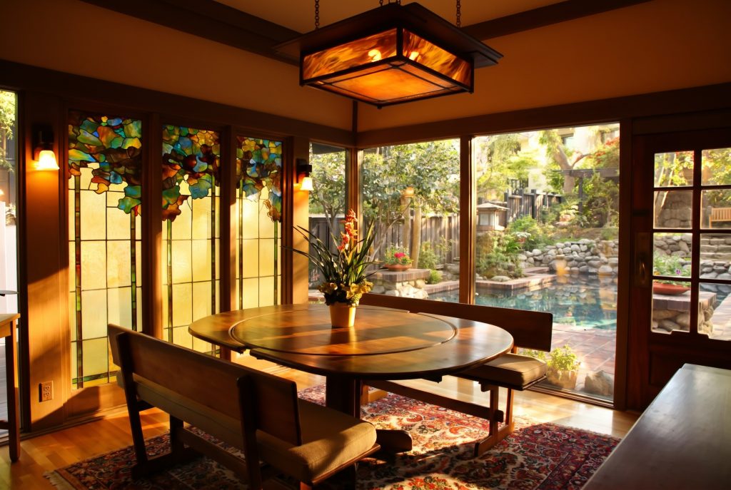 A breakfast nook lit by a hanging amber art-glass lantern, with three tall leaded-glass windows in a colorful foliage pattern beside a wall of windows looking onto a koi pond and garden.