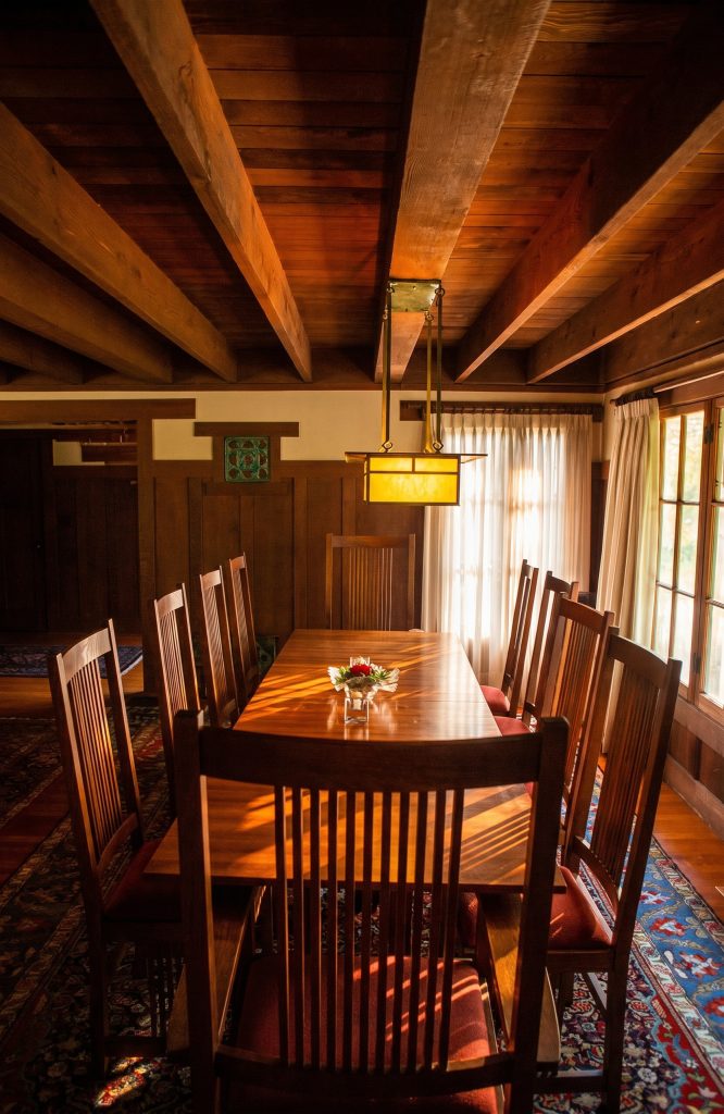A wood-paneled dining room with a low beamed ceiling, a long table and spindle-back chairs, and an amber art-glass lantern suspended on brass straps; a green glazed tile is set into the frieze at left.
