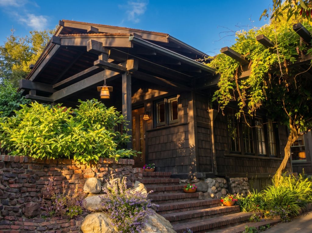 The deep gabled entry porch of the shingled house at golden hour, exposed beams and a hanging lantern over brick steps that rise past river boulders and flowering plants.