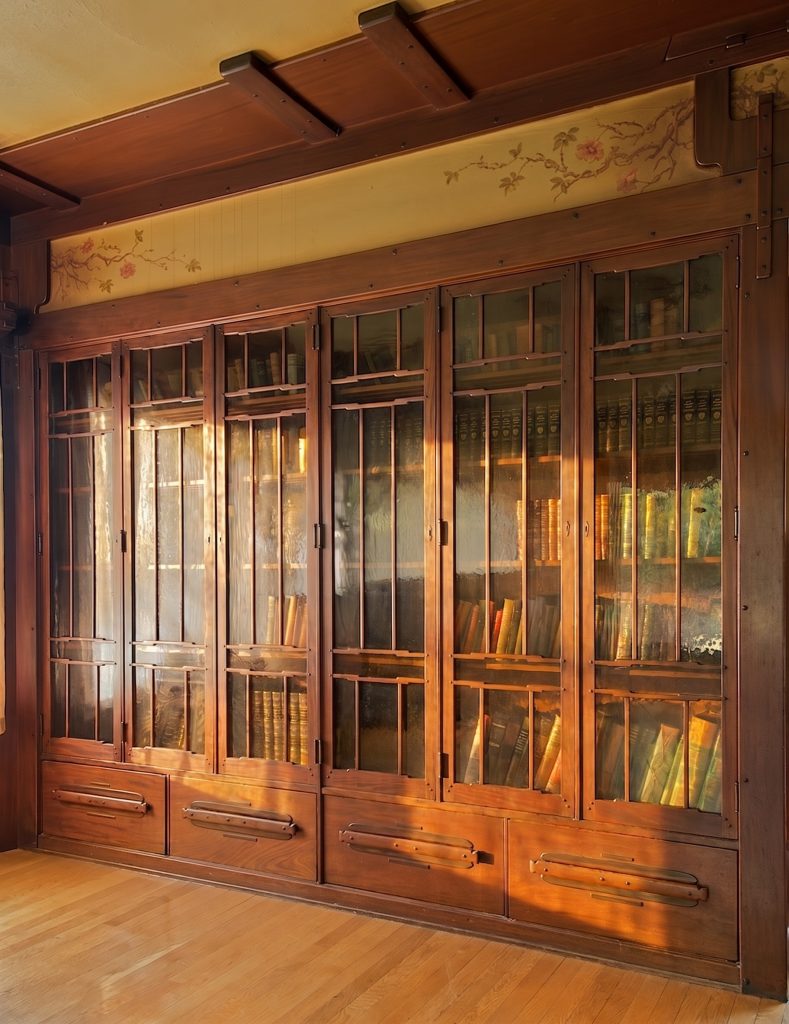 A long run of built-in mahogany bookcases with glazed doors and stepped muntins, deep drawers below with rounded pulls and square pegs, and above them a hand-painted frieze of wild roses on tan linen, beneath a heavy pegged ceiling beam.