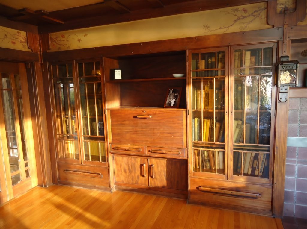 A built-in mahogany cabinet wall in the Thorsen House: glass-doored bookcases full of leather-bound volumes flanking a central drop-front desk over drawers and paired cabinet doors, a painted frieze of pink roses on a branch running above, a beamed ceiling with square ebony pegs, and an art-glass wall lantern at right.