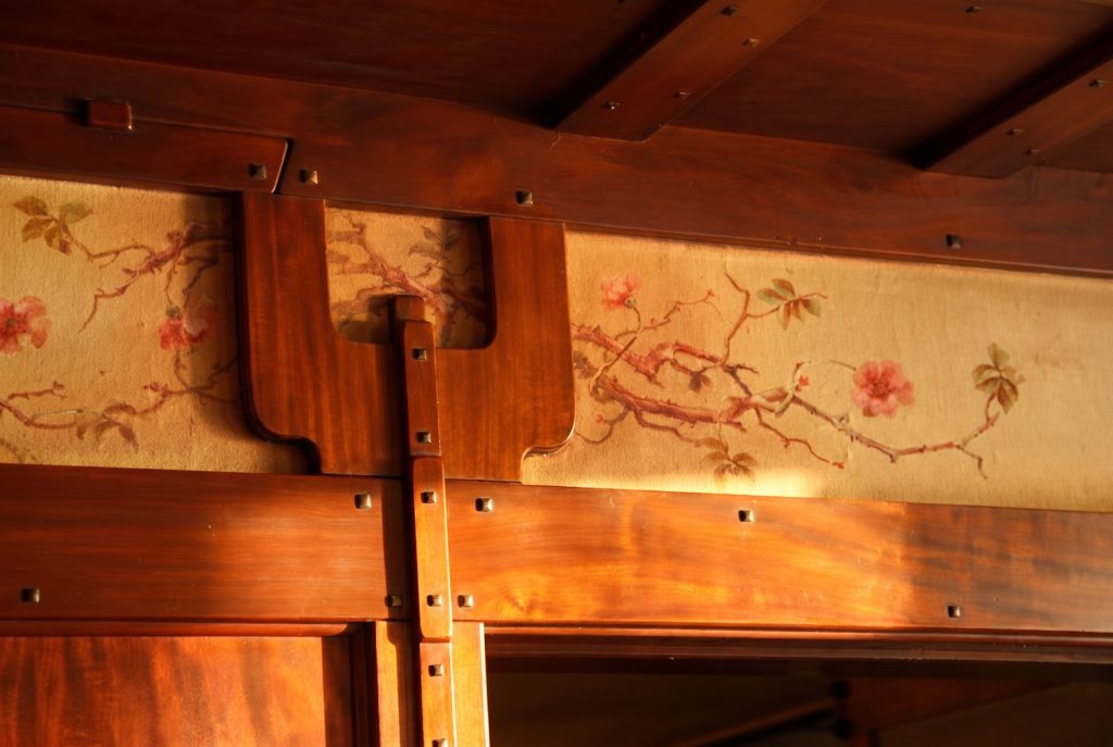 Close-up of the painted frieze: pink wild roses and buds on a thorned, leafy branch brushed onto a tan linen panel, set between mahogany members, with a vertical strap carrying a column of square ebony pegs.