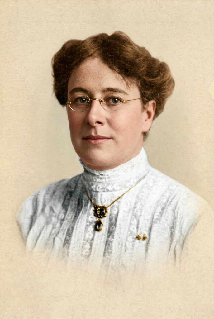 Colorized studio portrait of a woman with auburn upswept hair, gold oval pince-nez spectacles, and a white high-necked lace blouse with a gold pendant.