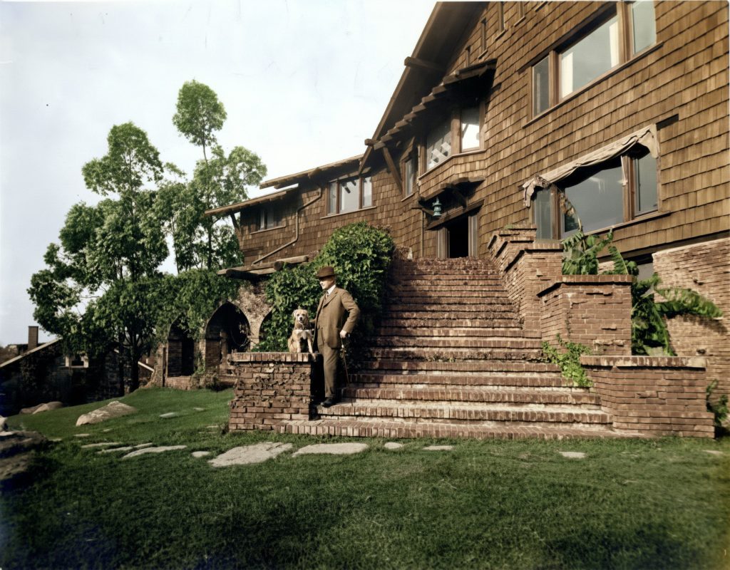 Nathan Bentz, in a three-piece suit and bowler hat and holding a cane, stands with a small dog on the broad clinker-brick entrance stairs of his two-story shingled Craftsman house, deep overhanging eaves and exposed rafters above, arched brick openings and mature trees to the left, and a lawn with stepping stones in the foreground.