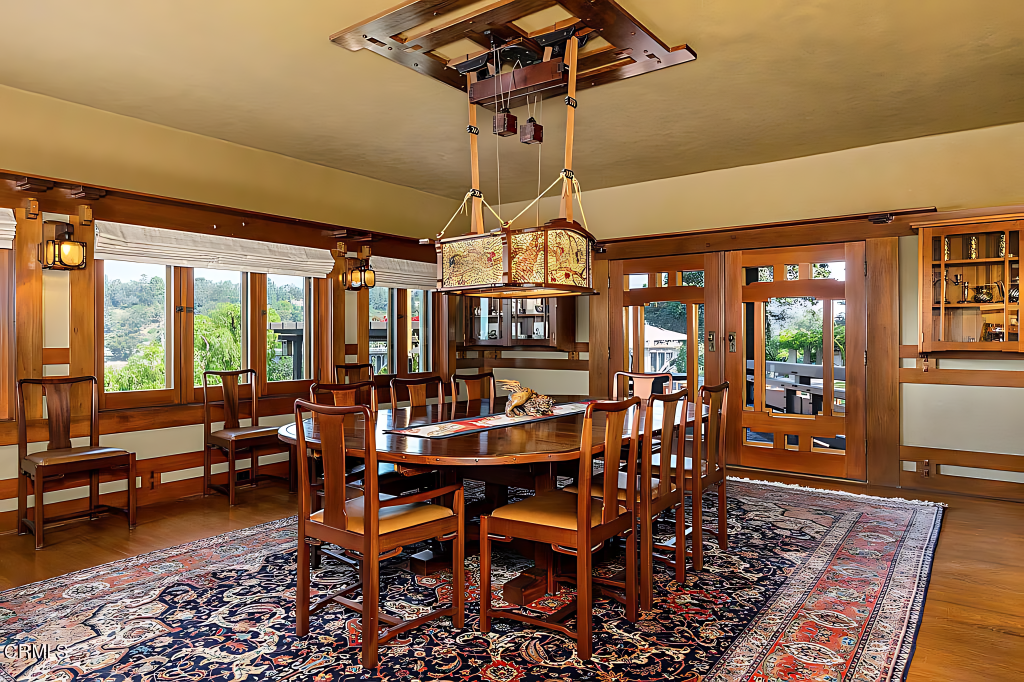 Sunlit dining room of the Robinson House in Pasadena today, furnished with reproduction Greene and Greene pieces — a long table ringed by chairs on a large Persian rug, a suspended leaded-glass fixture overhead, wood-framed windows and French doors looking out to a green canyon, with built-in cabinets and lantern sconces.