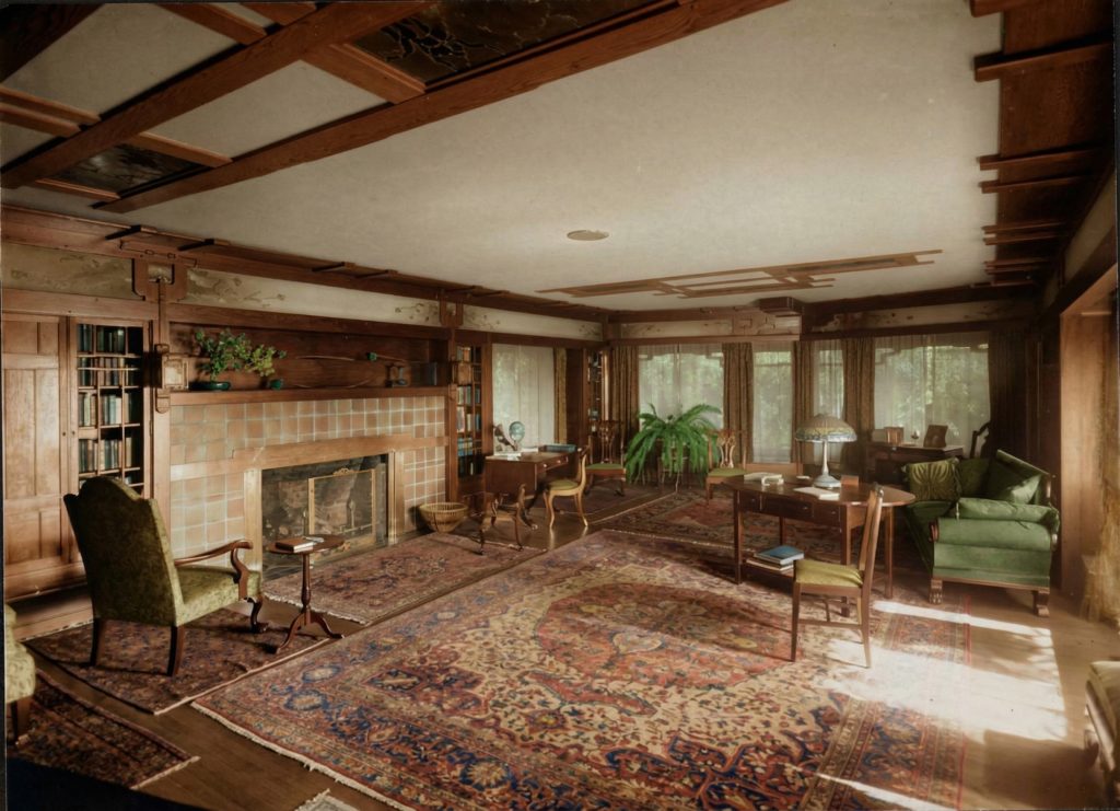 Historical photograph of the Thorsen House living room, the library table standing on a large patterned carpet between the tiled fireplace and a bank of curtained windows, with built-in bookcases, a green upholstered wing chair, and a beamed ceiling.