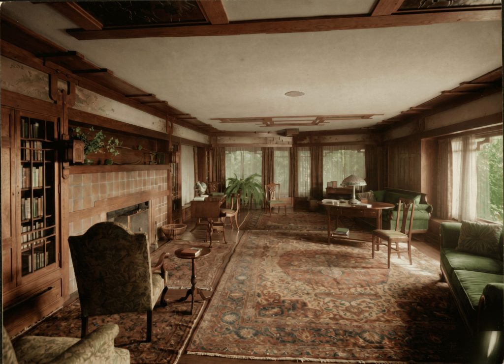 A rendered view of the Thorsen House living room from a slightly different angle, showing the library table, a green settee and chairs, a table lamp, the tiled fireplace, built-in bookcases, and curtained windows onto the trees.