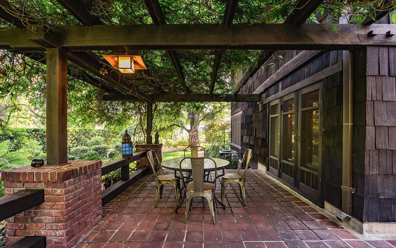 A brick-paved terrace at the Louise Bentz House sheltered by a wisteria-covered pergola, with a small table and chairs beside the shingled house wall.