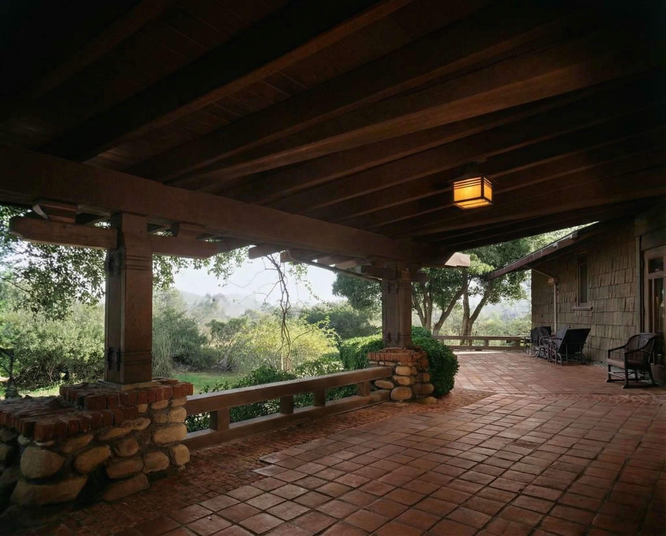 The deep covered rear terrace of the Charles M. Pratt House, heavy timber beams on boulder-and-brick piers, a box lantern hanging above the tile floor, valley beyond the low rail.